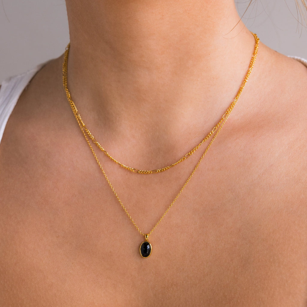 A woman styles the Onyx Figaro Duo Necklace—featuring two gold chains and a bold black onyx pendant—over bare skin and a white top.