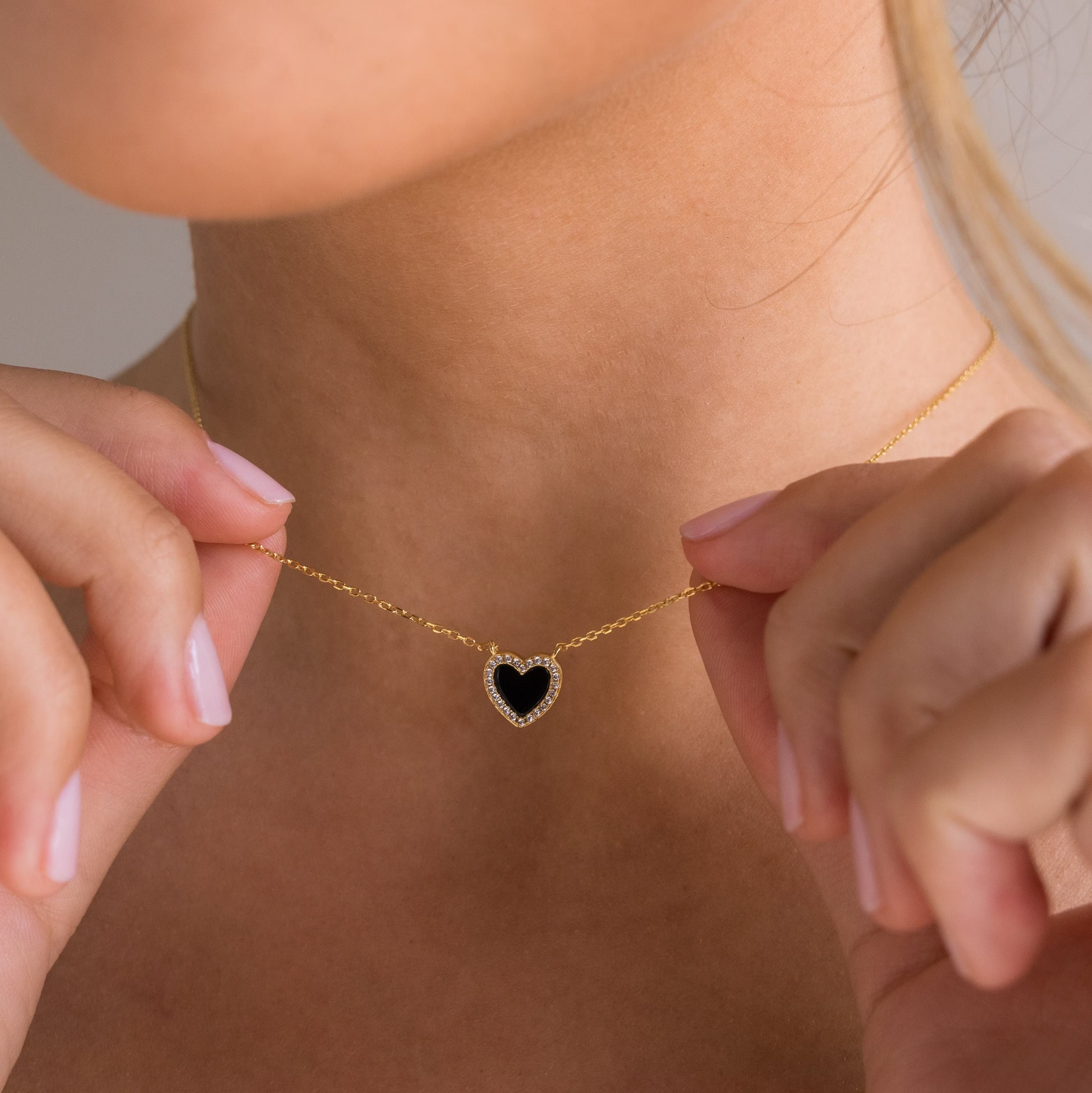 A woman holds the Pave Onyx Heart Necklace close to her neck, its black heart-shaped charm outlined with sparkling crystals.