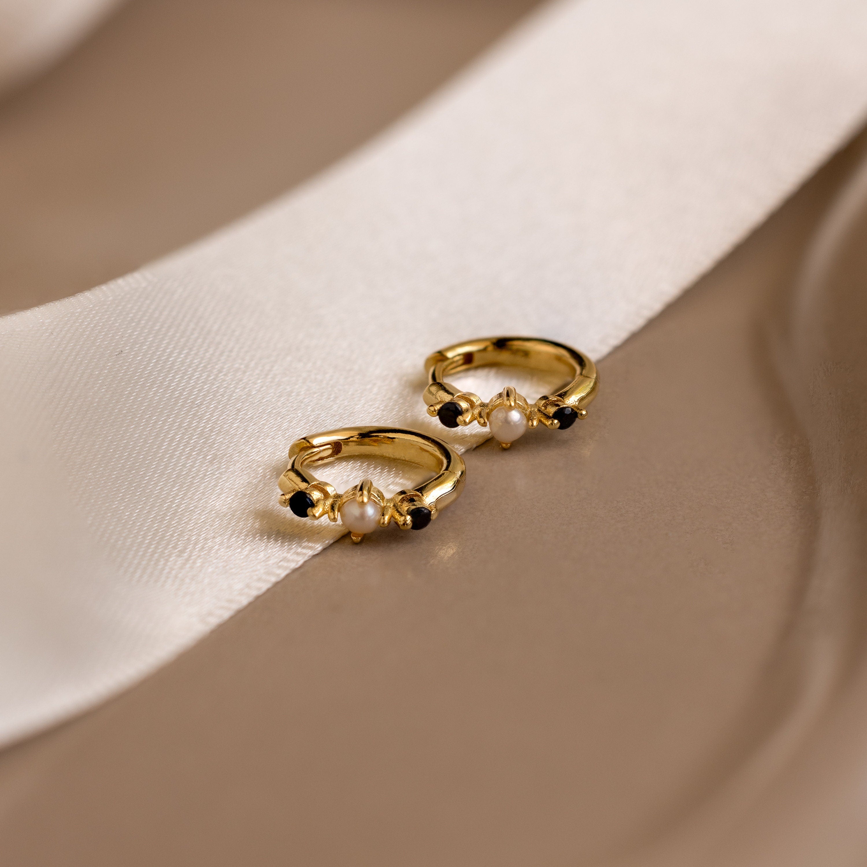 The Pearl & Onyx Huggies, featuring delicate pearl and black stone accents, rest on a beige surface beside a white ribbon.