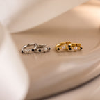 The Pearl & Onyx Huggies—one pair in silver, one in gold—feature pearls and black stones for a chic, elegant look on a beige surface, ideal for anyone seeking timeless pearl earrings.