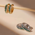 Two pairs of hoop earrings—one gold, one silver—sparkle with green and white stones on a beige surface. Get inspired by the Emerald Pave Snake Hoops, crafted to add bold style and elegance to any look.