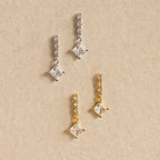 Two pairs of Princess Diamond Drop Studs—one in silver, one in gold—adorn a beige textured background, each pair featuring modern square crystals that highlight the sparkle of a princess cut gemstone.