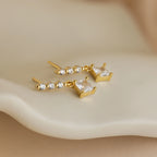 Princess Diamond Drop Studs featuring square princess cut gemstones and round clear stones, shown on a soft cream surface.