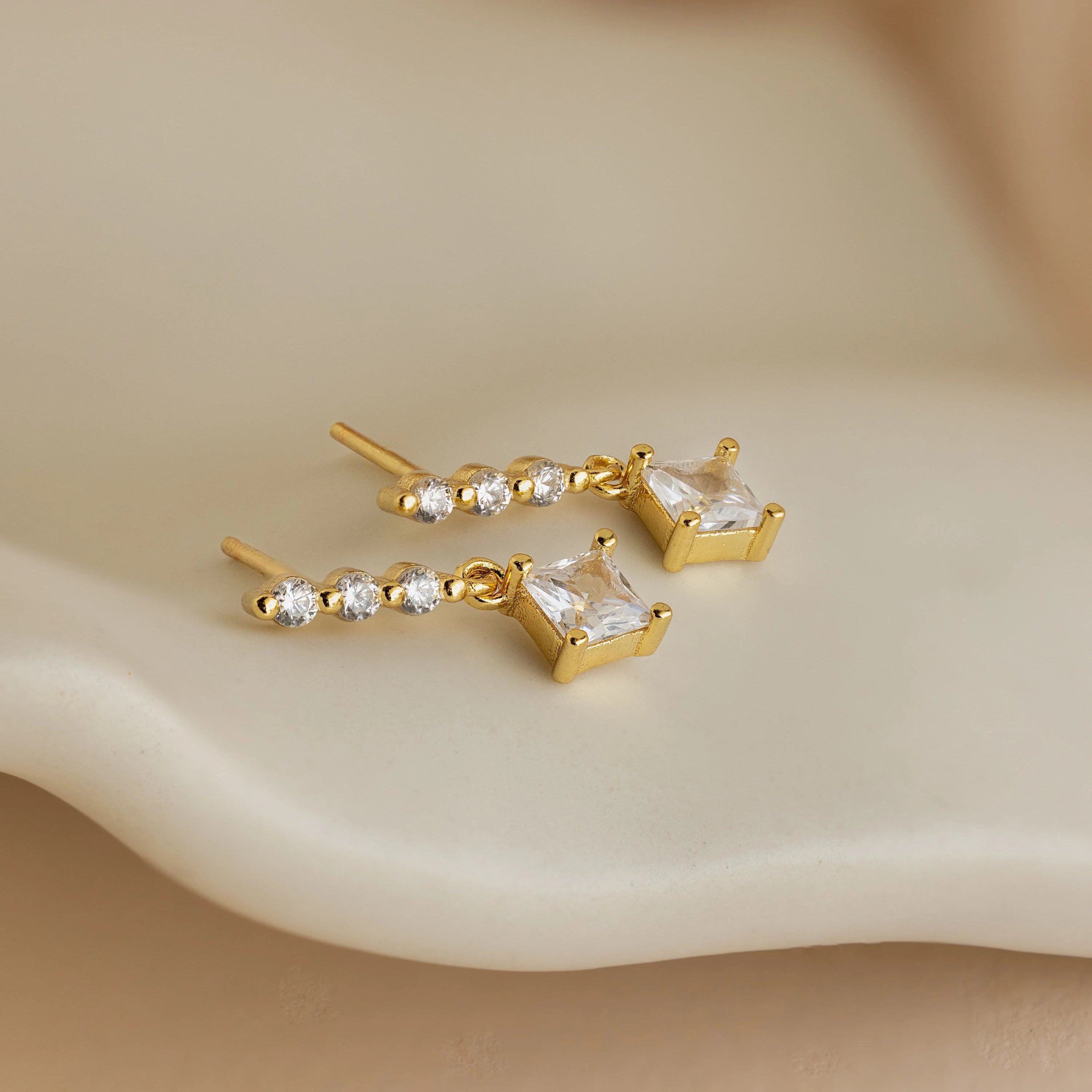 Princess Diamond Drop Studs featuring square princess cut gemstones and round clear stones, shown on a soft cream surface.