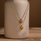 Two Diamond Ribbed Heart Necklaces, one in silver and one in gold, hang on chains draped over a neutral ceramic vase.
