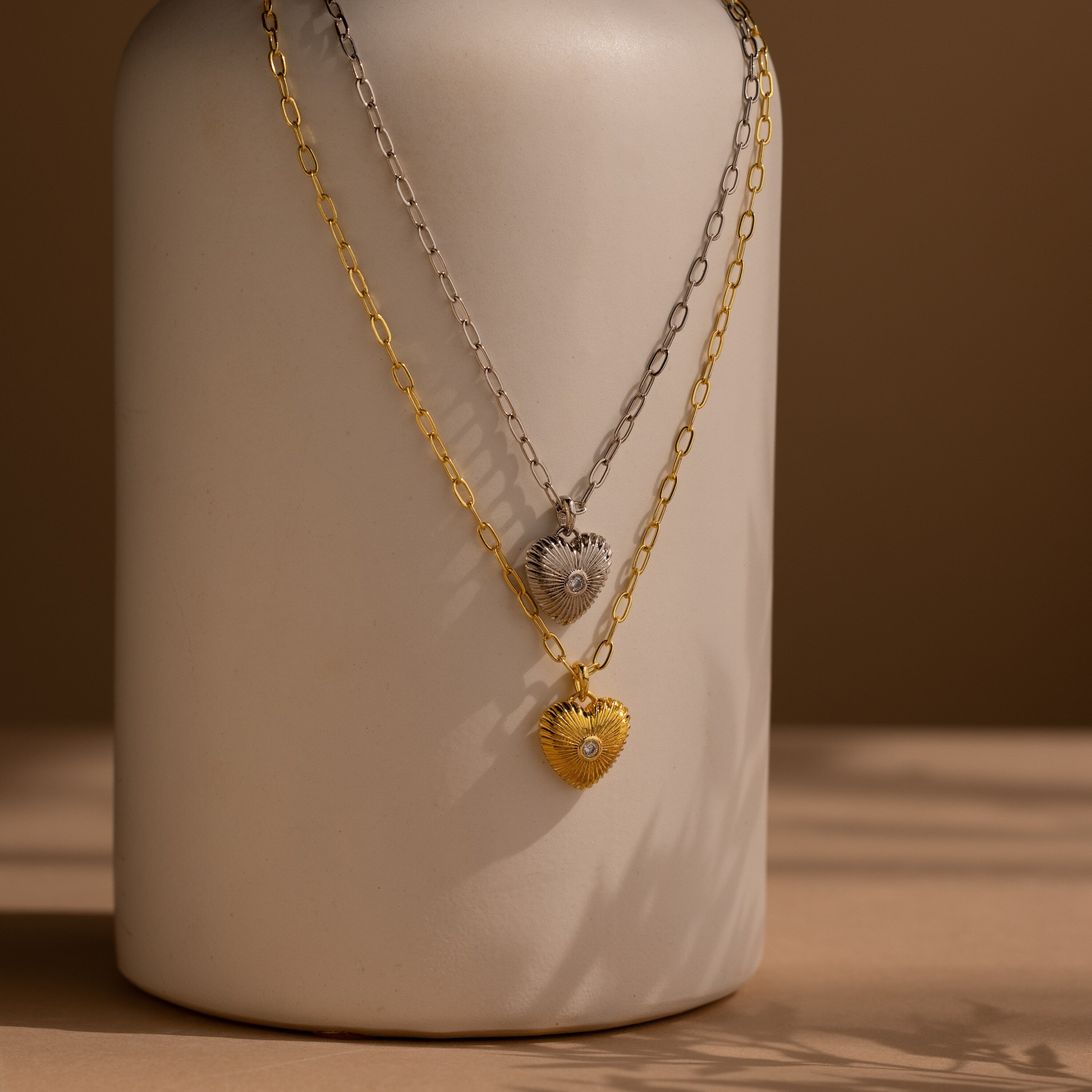 Two Diamond Ribbed Heart Necklaces, one in silver and one in gold, hang on chains draped over a neutral ceramic vase.