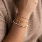 A person wears three elegant gold and diamond bracelets, including the dazzling 3 Prong Tennis Bracelet, on their wrist while resting on a beige sweater.