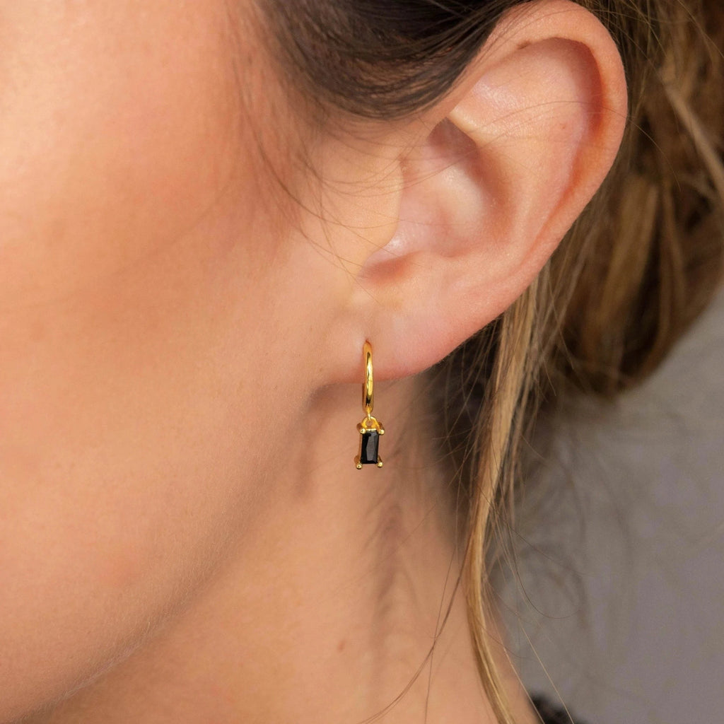 A close-up of an ear wearing the Onyx Baguette Drop Huggies, gold hoops adorned with a small rectangular black onyx stone.