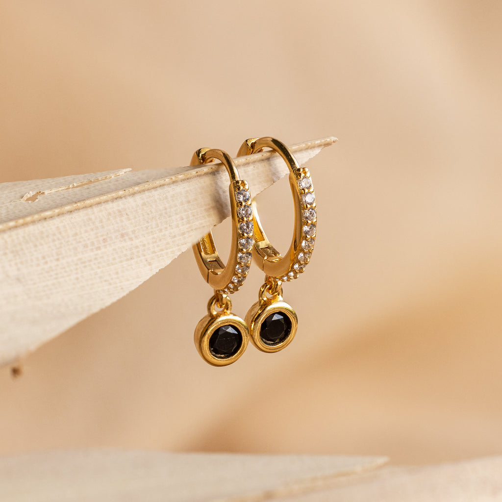 The Pave Onyx Drop Huggies showcase sparkling pavé stones and dangling black Onyx charms, beautifully displayed on a beige surface.
