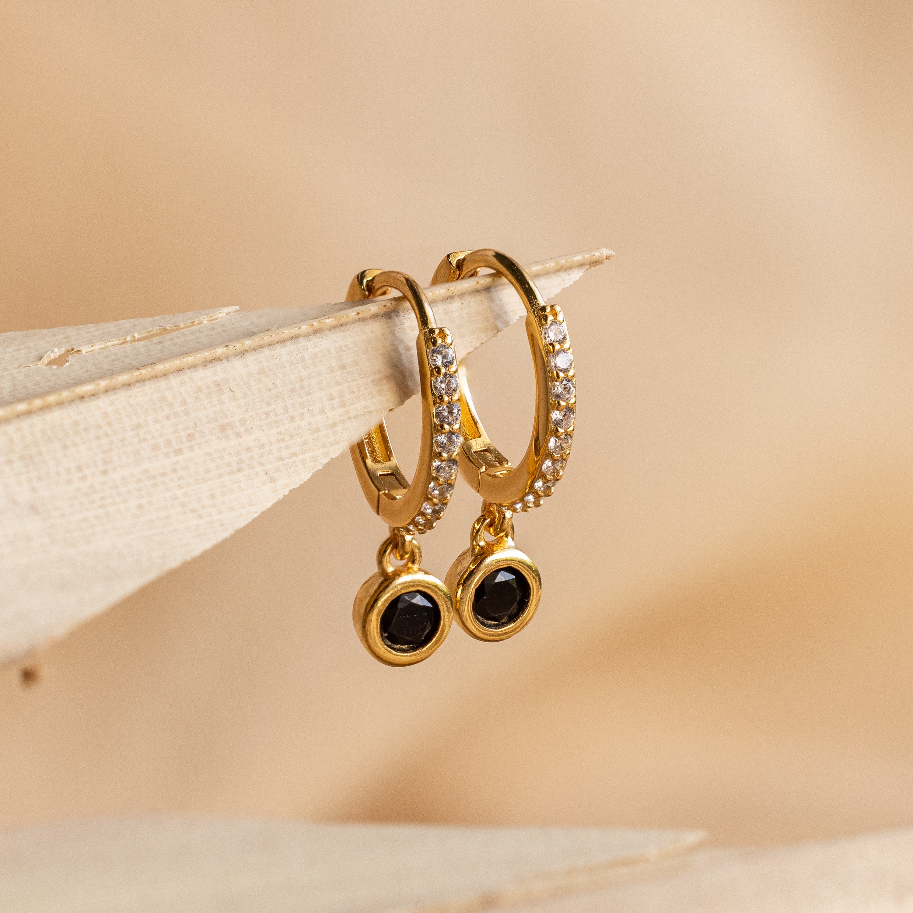 The Pave Onyx Drop Huggies showcase sparkling pavé stones and dangling black Onyx charms, beautifully displayed on a beige surface.
