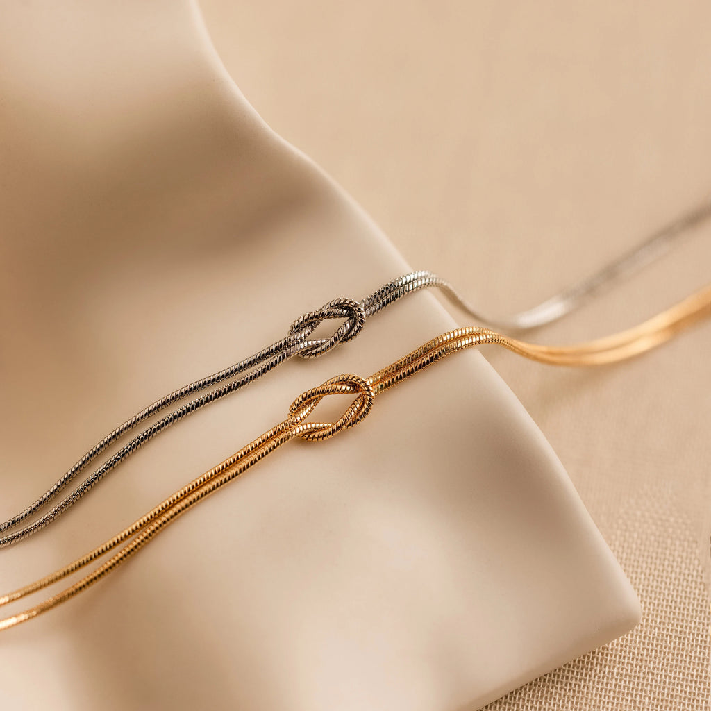 A pair of Love Knot Bracelets in white gold, each featuring a knot detail and double chain, elegantly displayed on a soft beige surface—an ideal gift for couples.