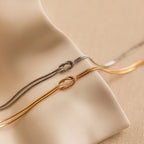 A pair of Love Knot Bracelets in white gold, each featuring a knot detail and double chain, elegantly displayed on a soft beige surface—an ideal gift for couples.