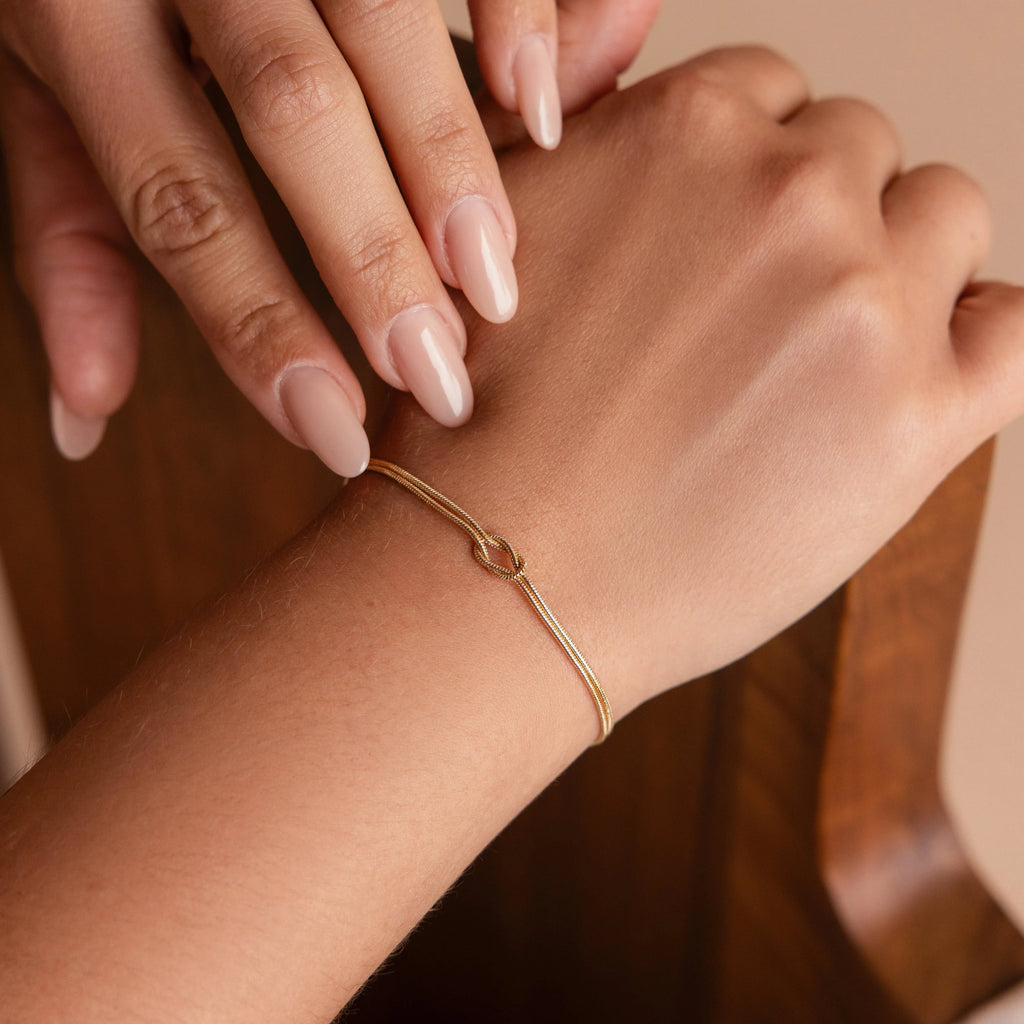 A hand with nude nails wears the Love Knot Bracelet in 18K Gold, featuring a small knot detail and elegant double chains—a thoughtful gift for couples.