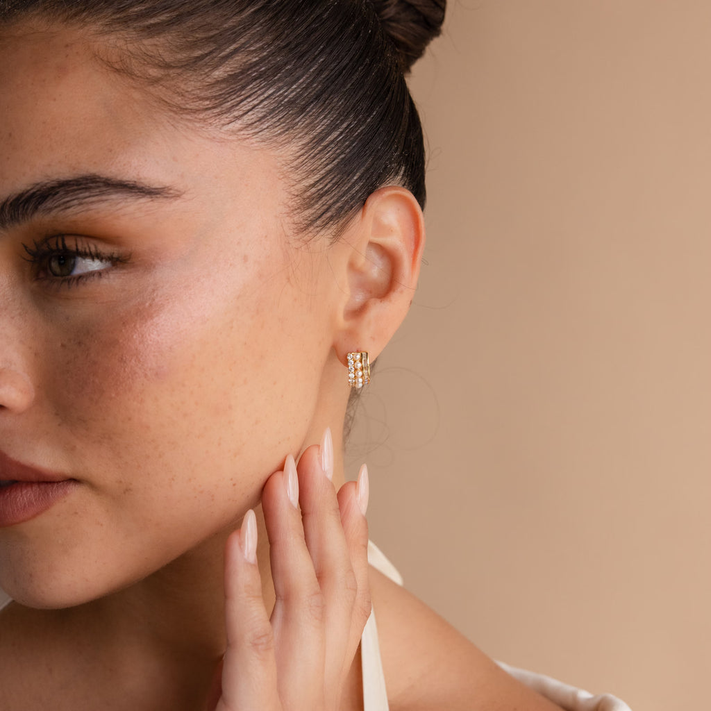 A woman with dark hair in a bun touches her face, wearing elegant Diamond Pearl Stacked Hoops bridal jewelry against a beige background.