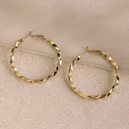 Large Twisted Hoops sit on a beige stone with light fabric draped above—an eye-catching summer accessory that adds effortless elegance.