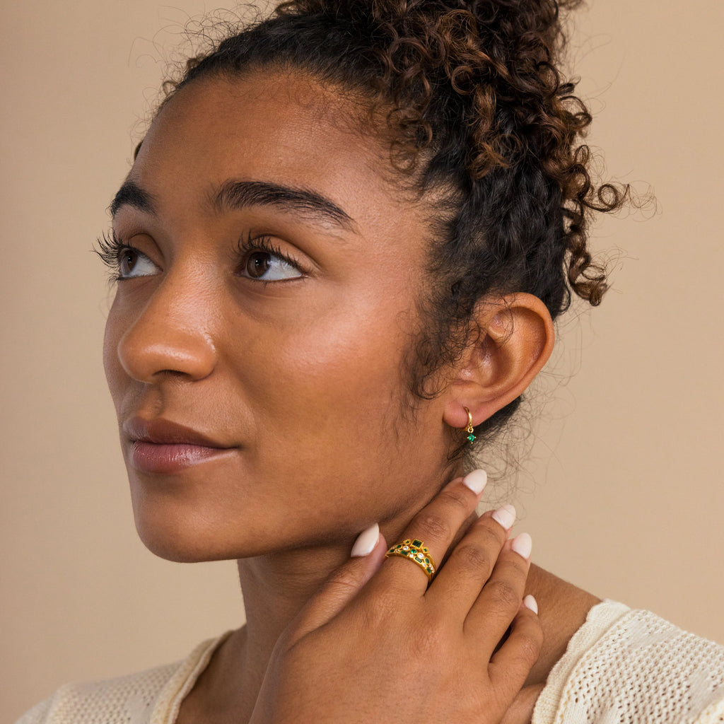 A woman with curly hair gazes to the side, hand on her neck, elegantly wearing gold Birthstone Drop Huggies and a cream top.