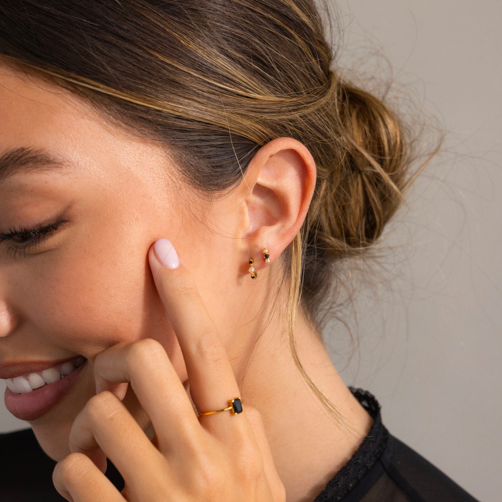 A woman smiles, touching her face and wearing Pearl & Onyx Huggies—double gold earrings with elegant pearl and dark stone details, complemented by a matching gold ring.