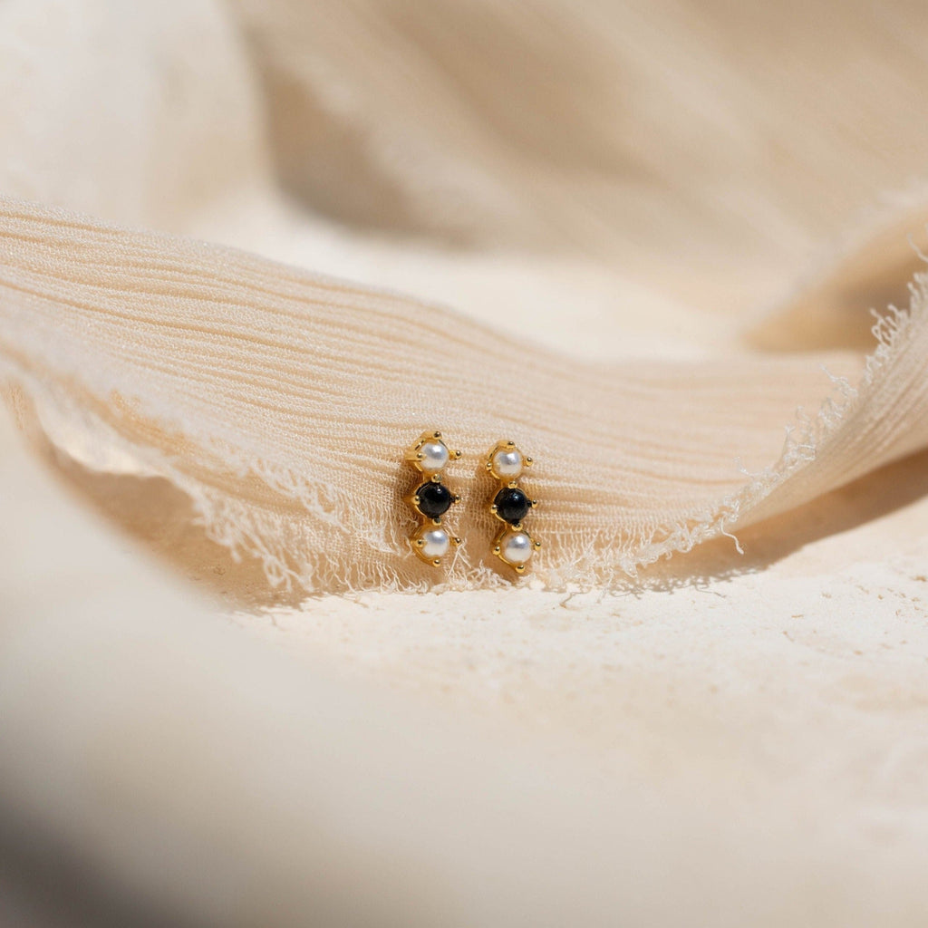 Tiny Onyx Pearl Bar Studs, featuring black stones and small pearls, are elegantly displayed on soft beige fabric for a chic look.