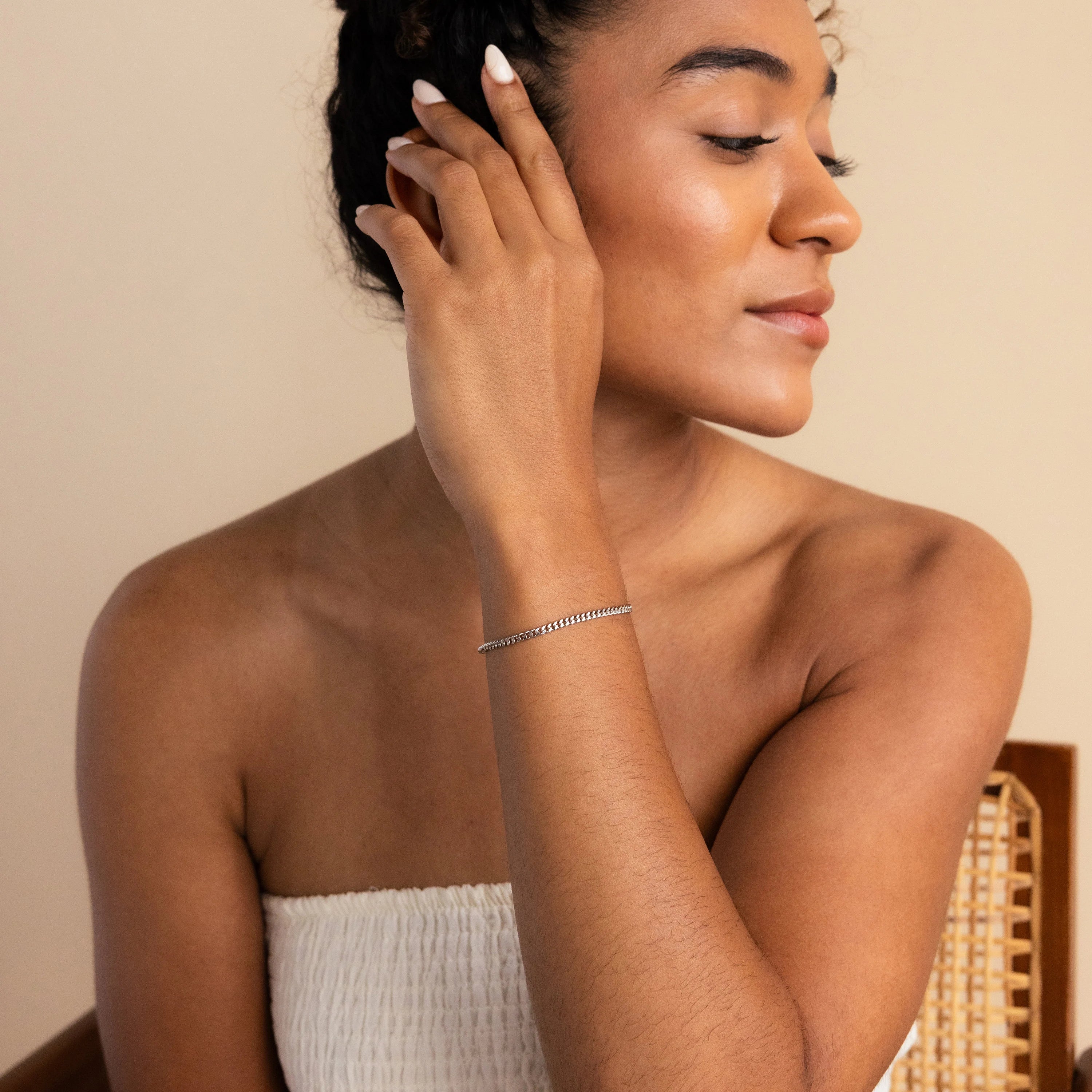 A woman in a white strapless top touches her ear, highlighting the minimal elegance of the Curb Chain Bracelet as she sits on a chair.