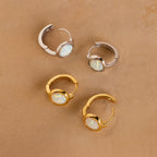 Four Opal Signet Huggies—two gold, two silver with round opals—are displayed on a tan textured surface, ideal for lovers of dainty hoops and understated style.