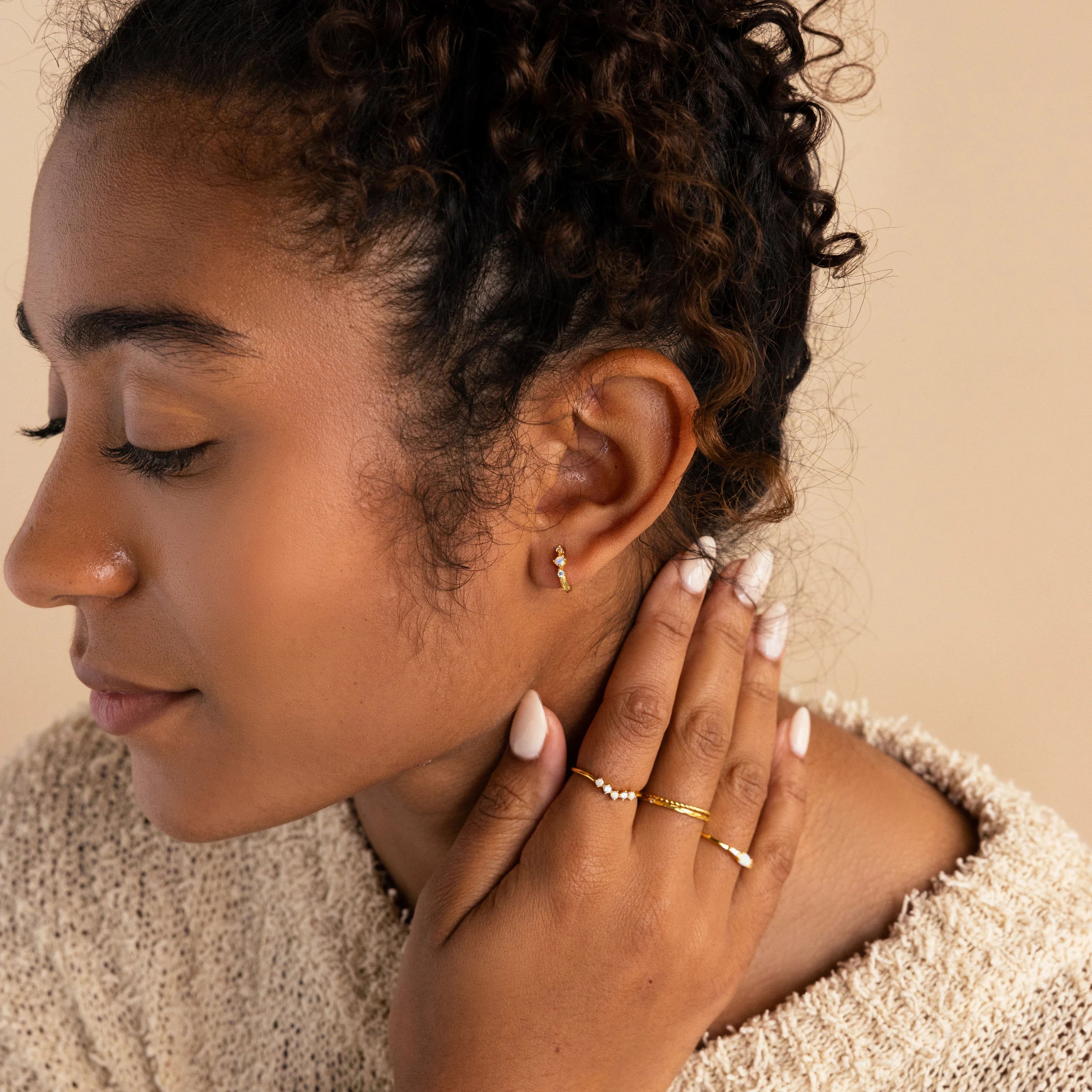 A serene portrait of a woman with her eyes closed and one hand resting on her neck, wearing a single gold opal and diamond huggie earring complemented by minimalist gold rings, creating a warm, cohesive jewelry look.