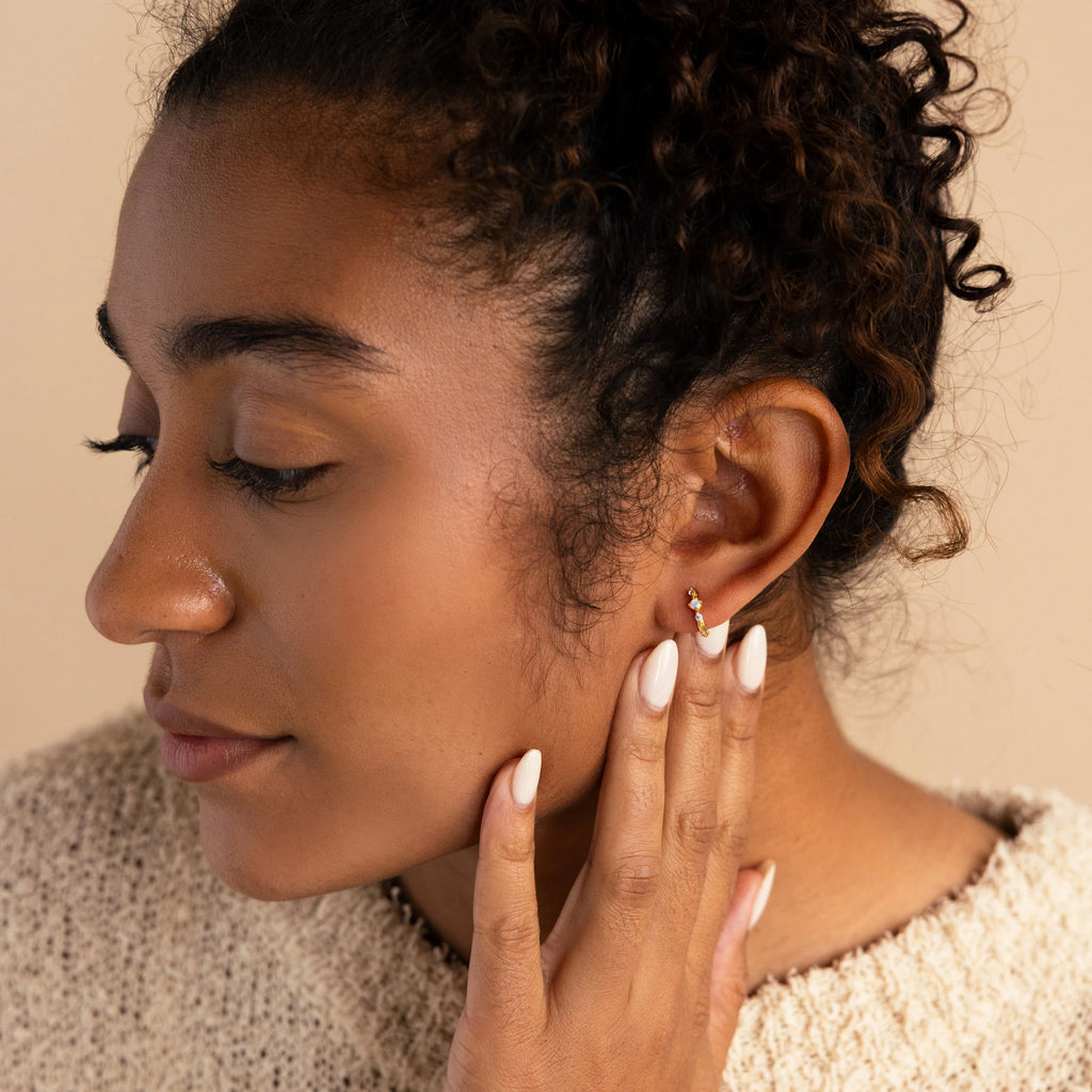 A side profile of a woman gently touching her ear, wearing a single gold opal and diamond huggie earring, styled with a cozy textured sweater