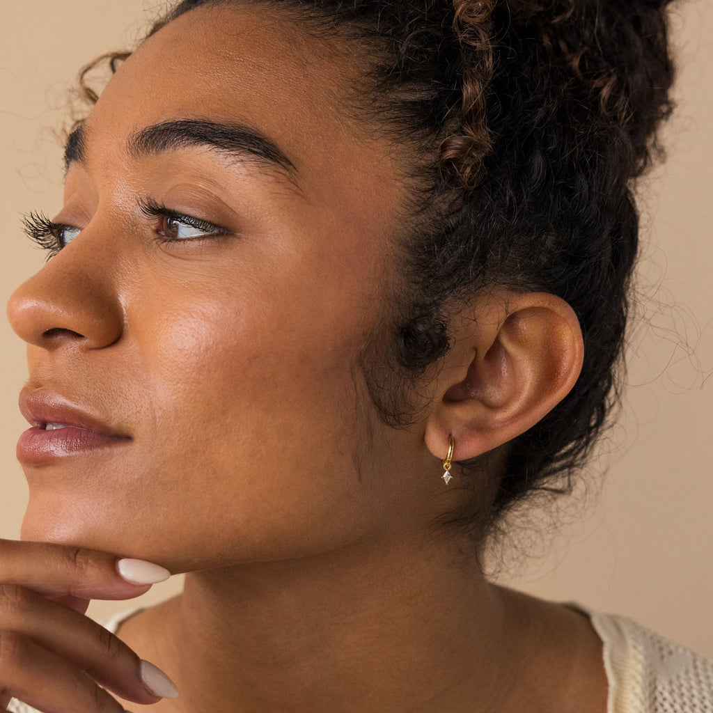 A woman thoughtfully admires her sparkling Birthstone Drop Huggies with her hand on her chin.