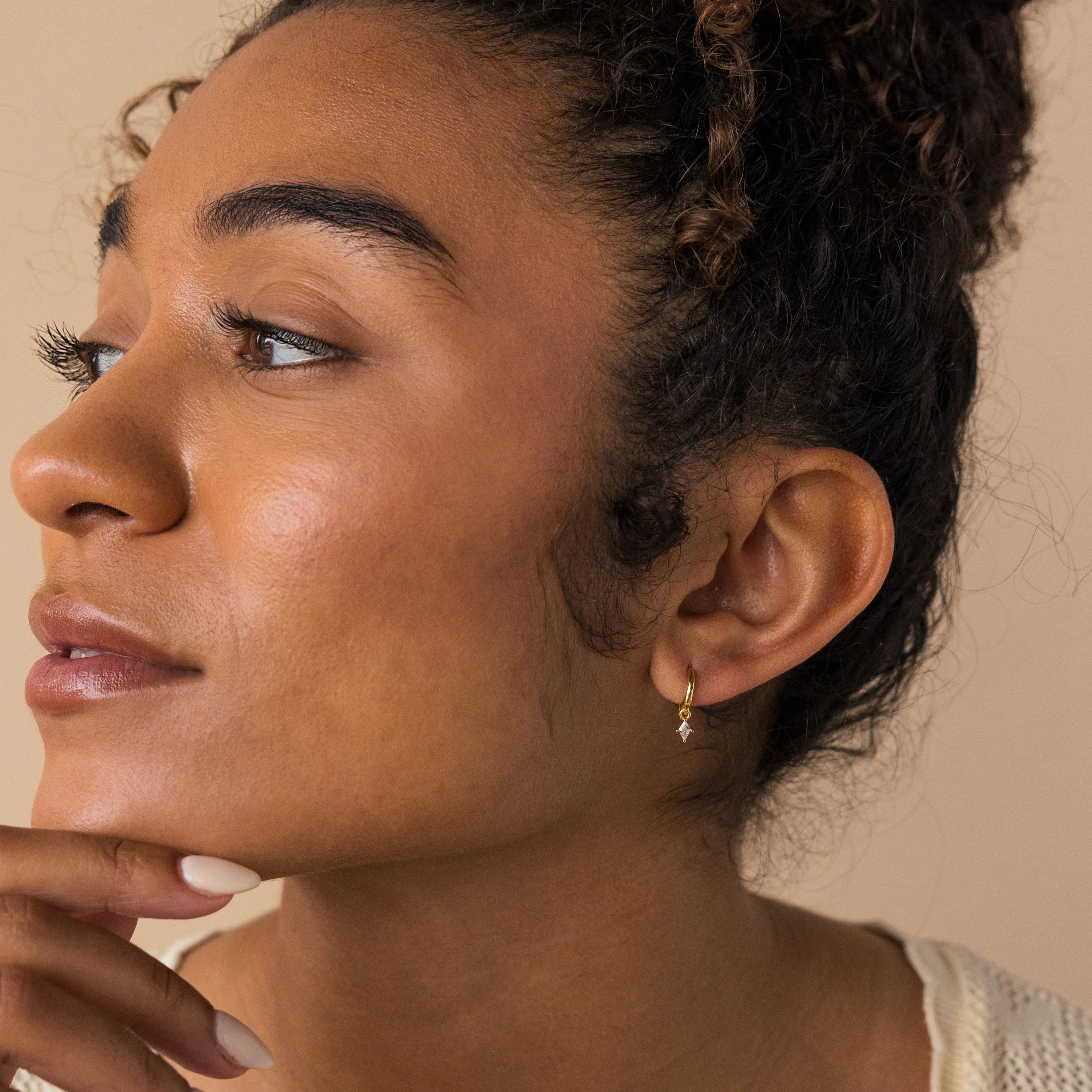 A woman thoughtfully admires her sparkling Birthstone Drop Huggies with her hand on her chin.