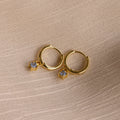 Birthstone Drop Huggies—gold huggie earrings featuring small square gemstone pendants, shown on textured beige fabric. Ideal as birthstone earrings or a meaningful piece of personalized jewelry.