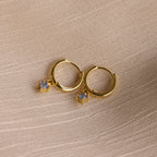 Birthstone Drop Huggies—gold huggie earrings featuring small square gemstone pendants, shown on textured beige fabric. Ideal as birthstone earrings or a meaningful piece of personalized jewelry.