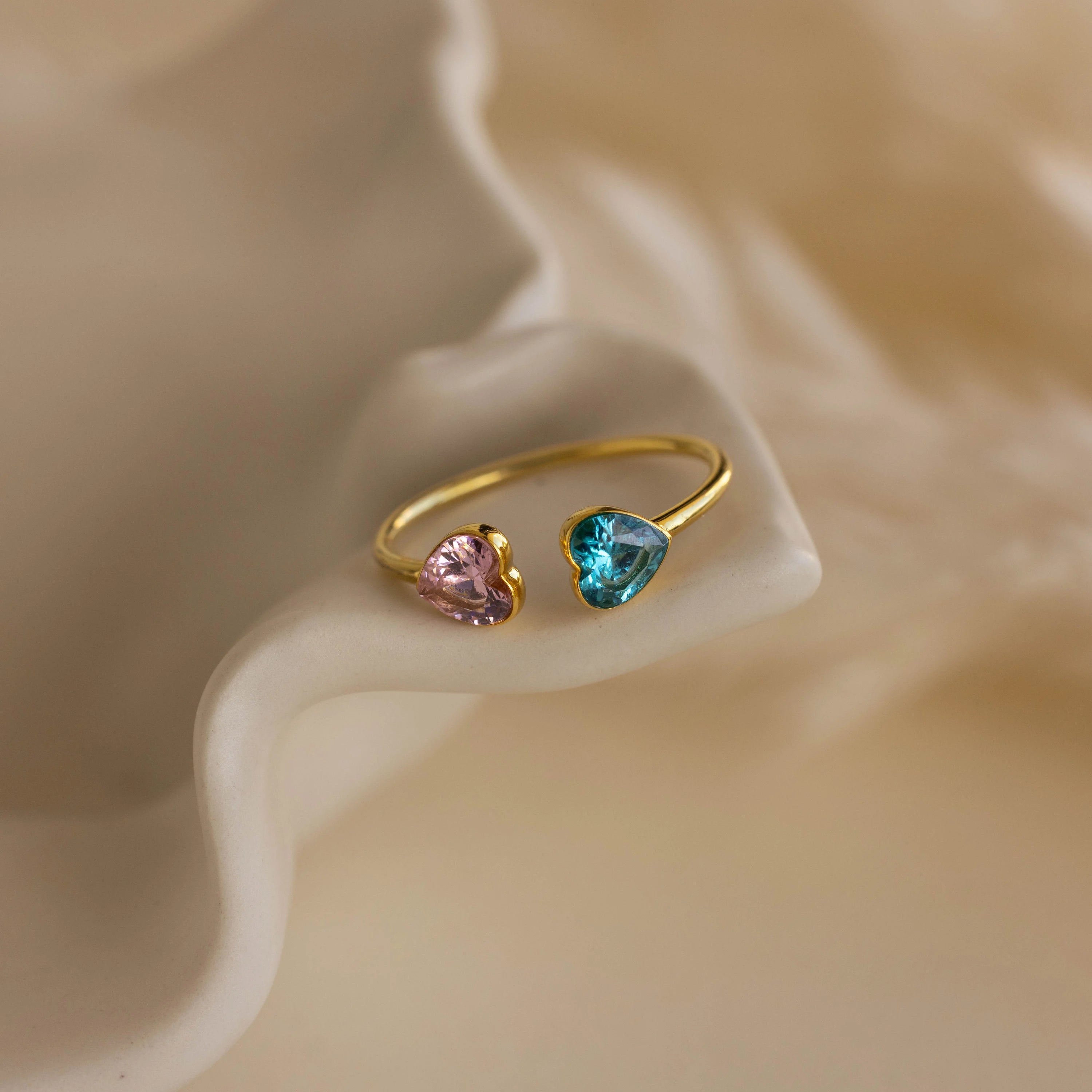 The Duo Heart Birthstone Ring, featuring pink and blue heart-shaped gems, is displayed on a wavy cream-colored surface.