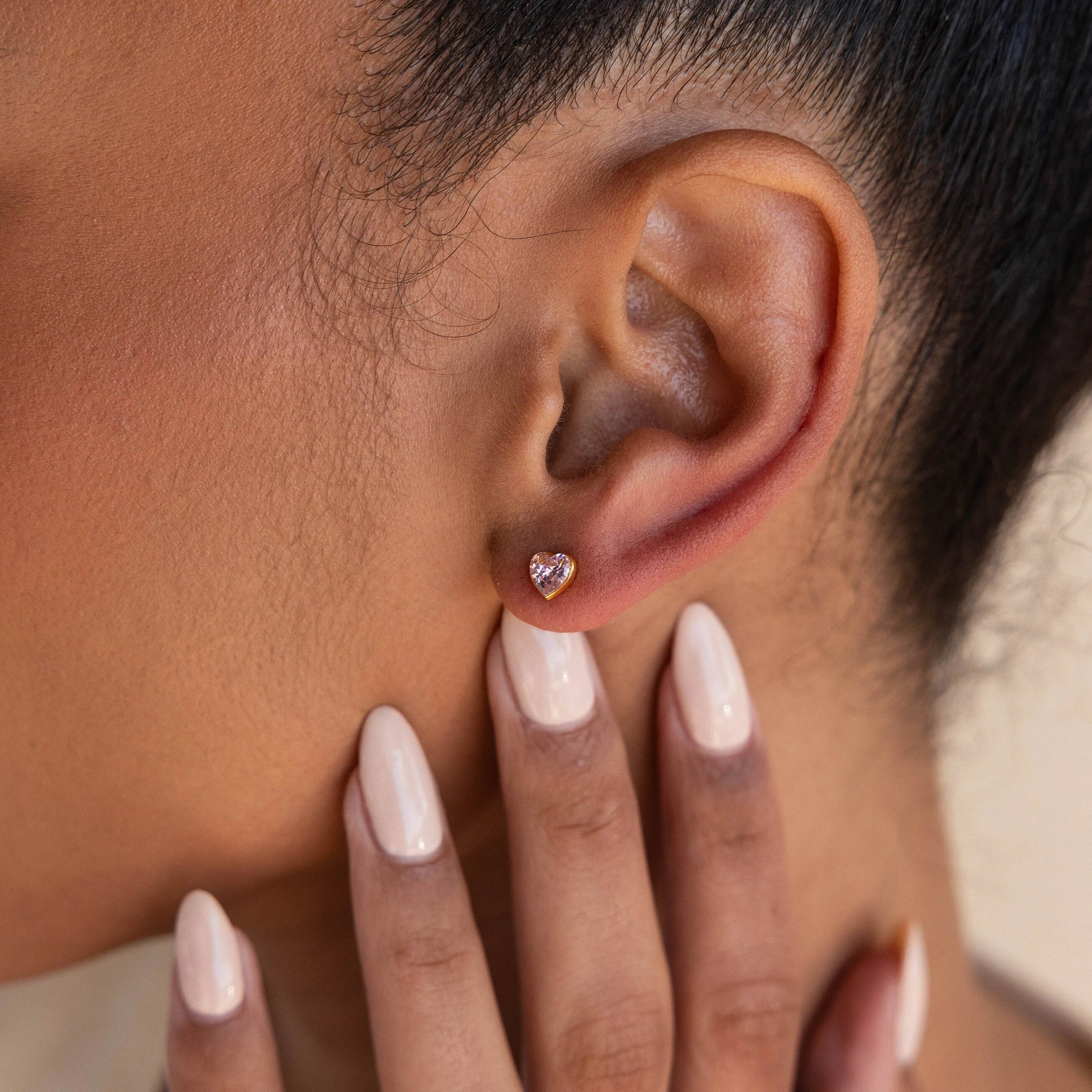 A manicured hand gently touches an ear adorned with Bezel Heart Birthstone Studs, highlighting the delicate heart-shaped gemstone earrings.