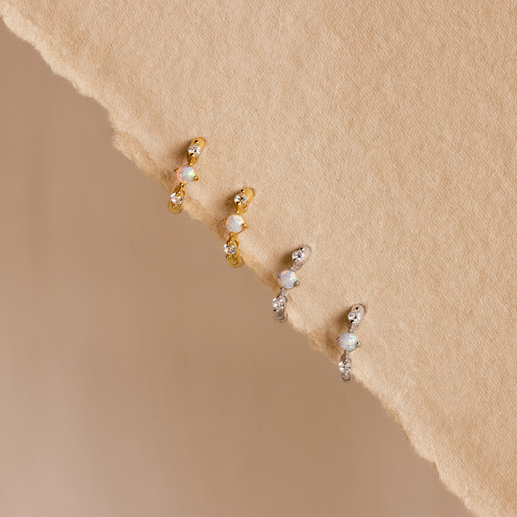 Four opal and diamond huggie earrings—two in gold and two in silver—neatly arranged on a textured beige background, highlighting the subtle sparkle and craftsmanship of each piece.