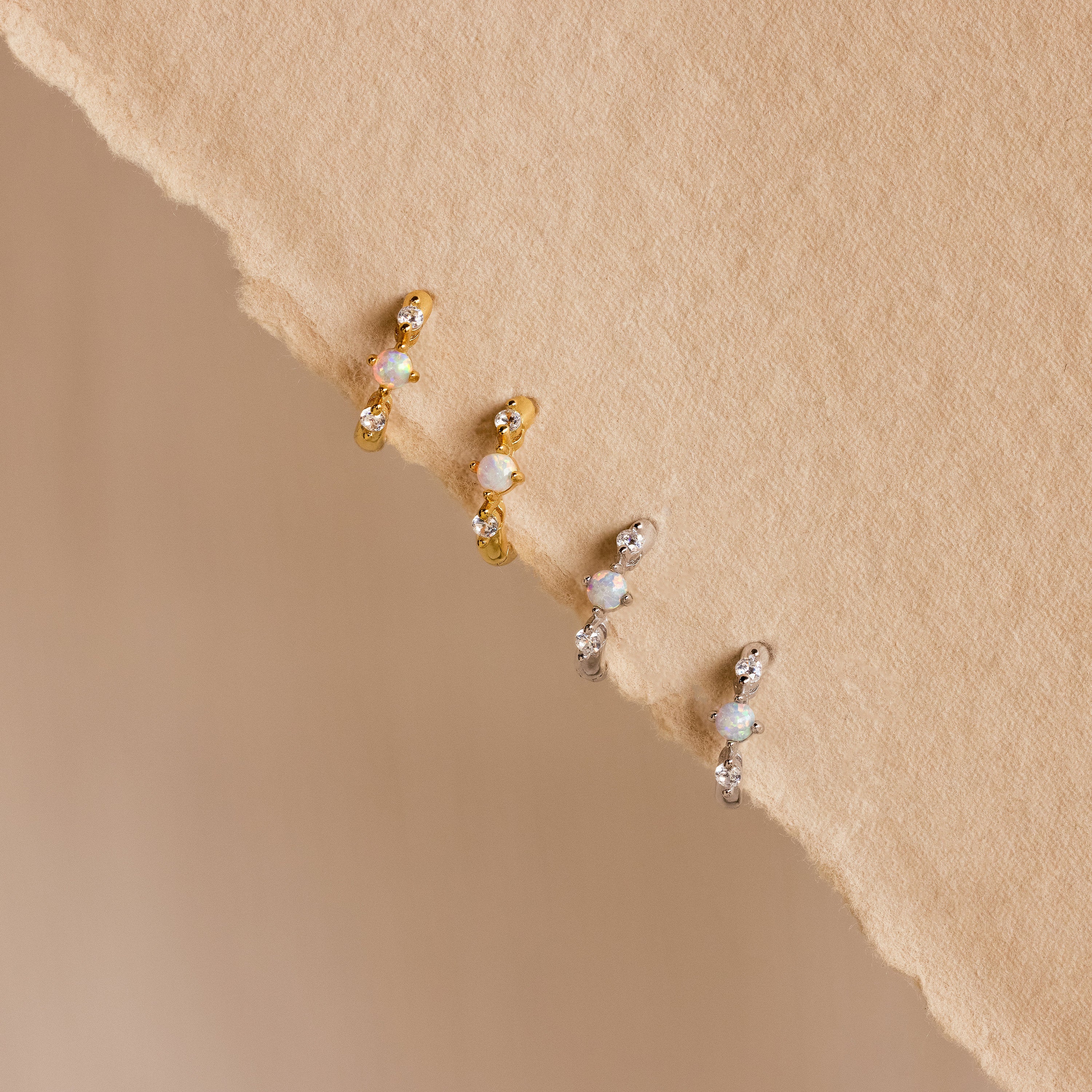 Four opal and diamond huggie earrings—two in gold and two in silver—neatly arranged on a textured beige background, highlighting the subtle sparkle and craftsmanship of each piece.