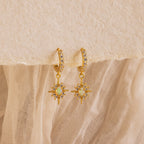 The Pave Opal Star Huggies capture celestial charm with gem-studded starburst pendants, beautifully showcased on a textured fabric background.