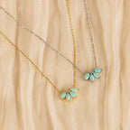 Two delicate necklaces rest on beige textured fabric: one features a gold floral design with a blue-green gemstone pendant, and the other is the Turquoise Opal Marquise Necklace in silver, both exuding understated elegance.