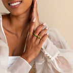 Woman in a sheer white blouse shows manicured nails and gold rings, highlighting the Ribbed Birthstone Signet Ring, while smiling and gently touching her face.