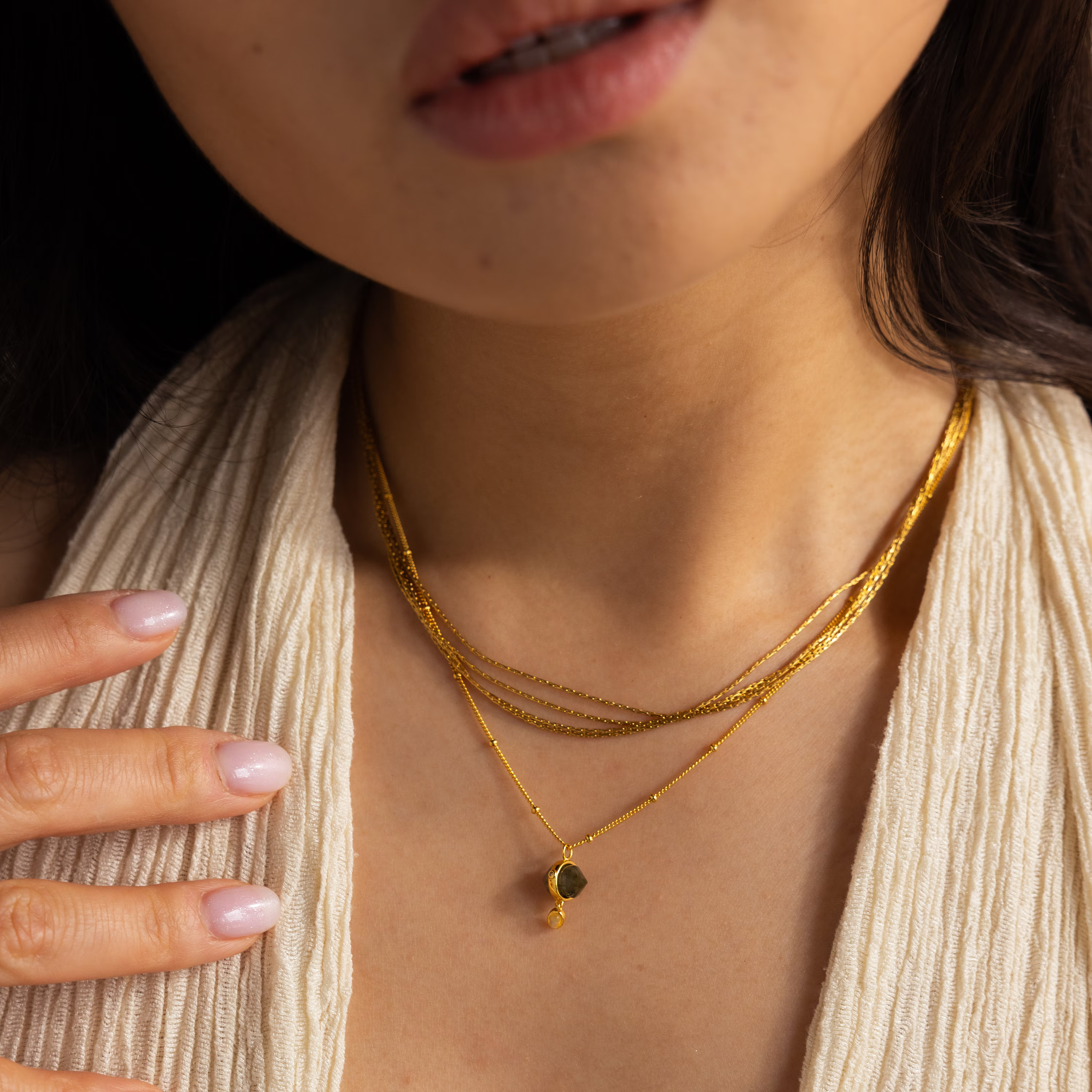 A woman wears layered gold necklaces, including the Labradorite & Moonstone Drop Necklace, with a cream textured top and her hand near her collarbone.