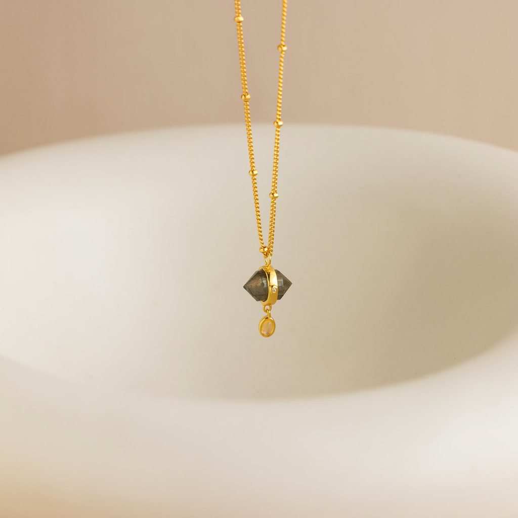 The Labradorite & Moonstone Drop Necklace features a shimmering gemstone pendant, suspended above a smooth, light-colored curved surface.