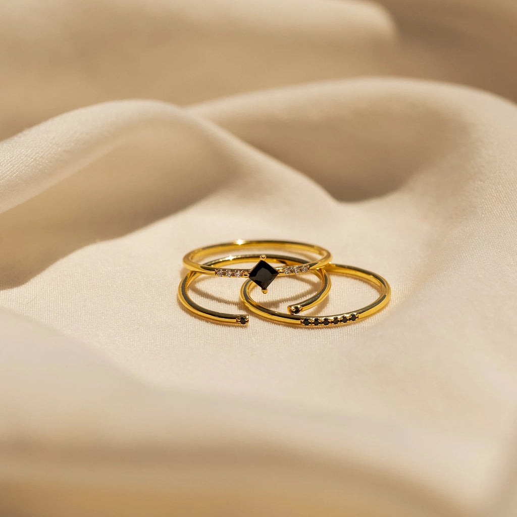 The Black Diamond Rings Set features three delicate rings with small black and clear stones, displayed on soft beige fabric—ideal for layering your jewelry.