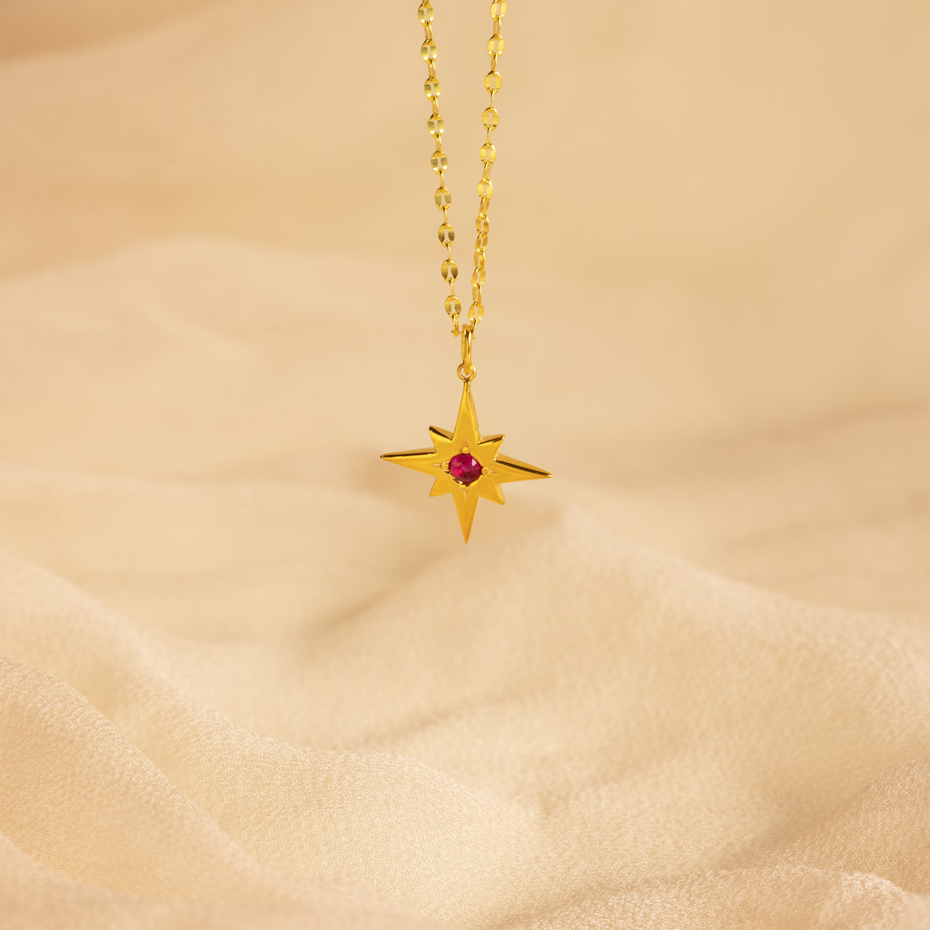 The Star Birthstone Necklace features a gold star-shaped pendant with a red gemstone on a chain, displayed on beige fabric—a radiant celestial jewelry piece and a meaningful personalized gift.