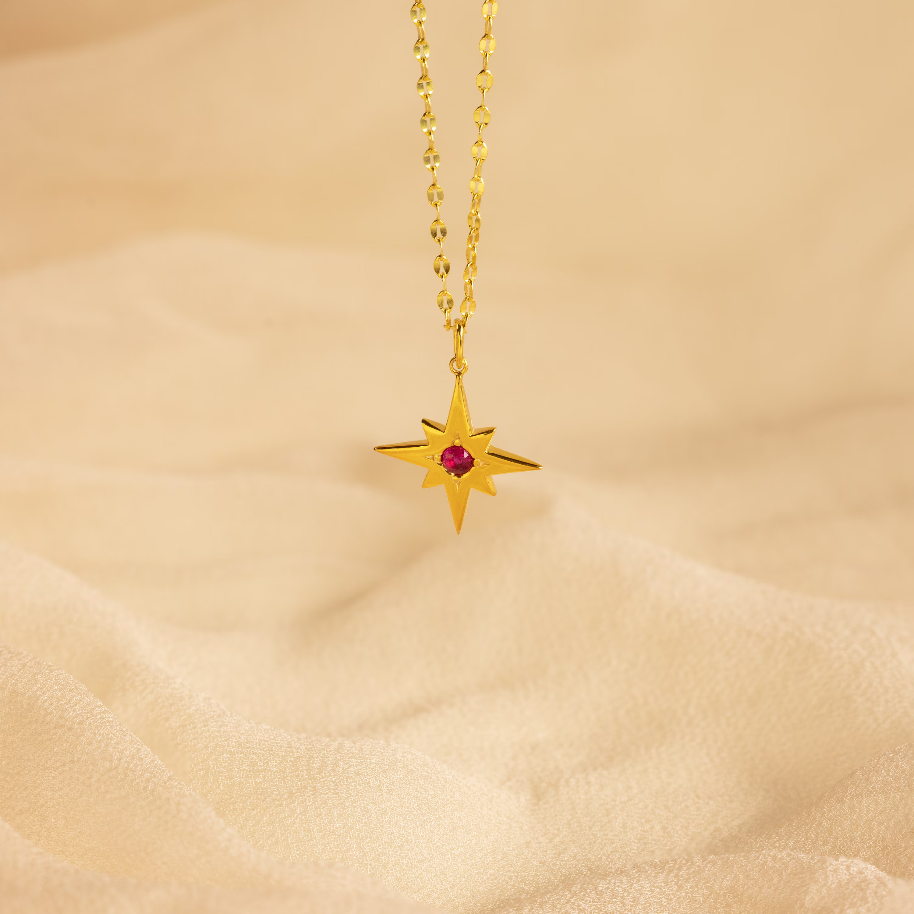 The Star Birthstone Necklace features a gold star-shaped pendant with a red gemstone on a chain, displayed on beige fabric—a radiant celestial jewelry piece and a meaningful personalized gift.