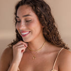 A smiling woman with curly hair wears the Opal Bezel Tennis Necklace layered with gold chains and a beige top, set against a neutral background.