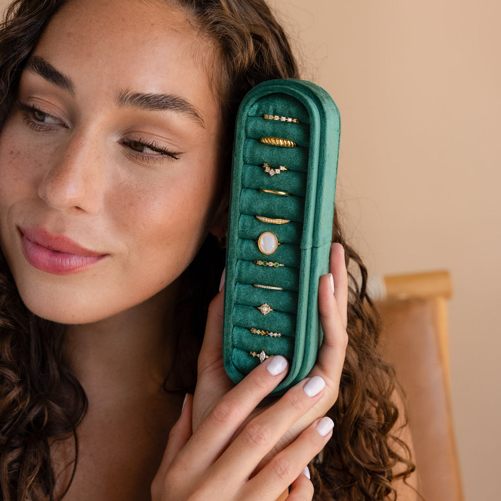 A woman smiles softly while holding the Slim Velvet Ring Box in green, displaying elegant gold rings.