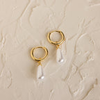 Pearl Teardrop Huggies: Gold hoop earrings with dangling teardrop-shaped pearls on a textured cream background, ideal for beach weddings or as elegant accessories for your special day.