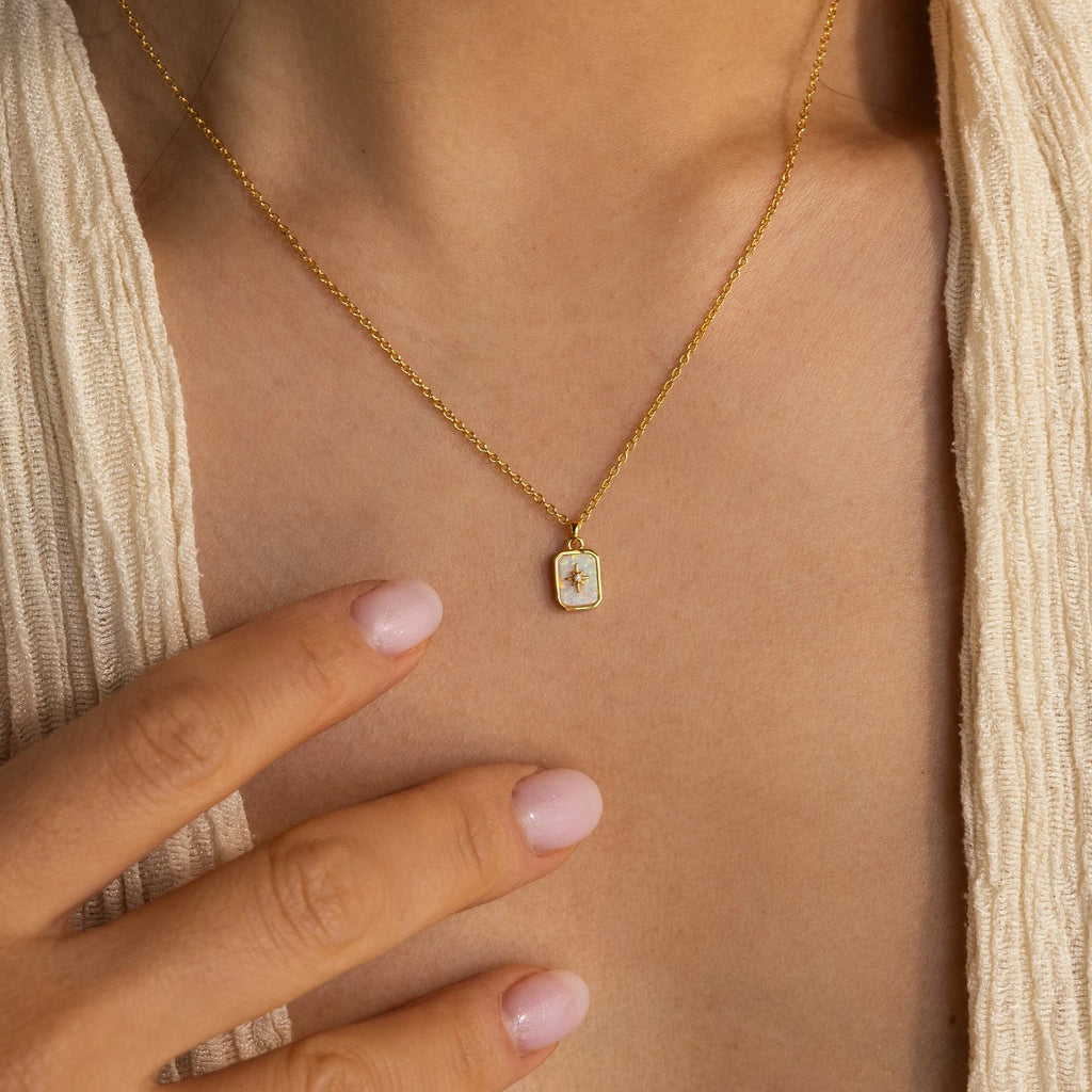 A woman wearing a beige textured top elegantly shows off the North Star Birthstone Necklace, which rests gracefully on her neck as her hand touches her chest.