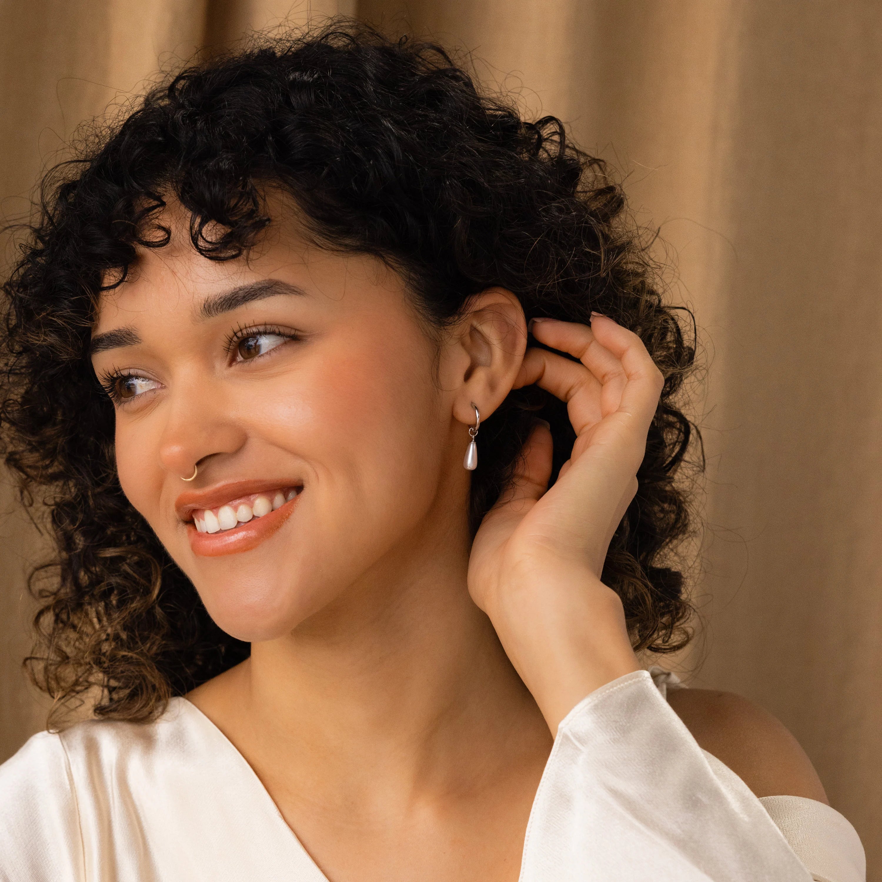 A smiling woman with curly hair wears Pearl Teardrop Huggies and a white top, touching her earrings in front of a beige curtain.