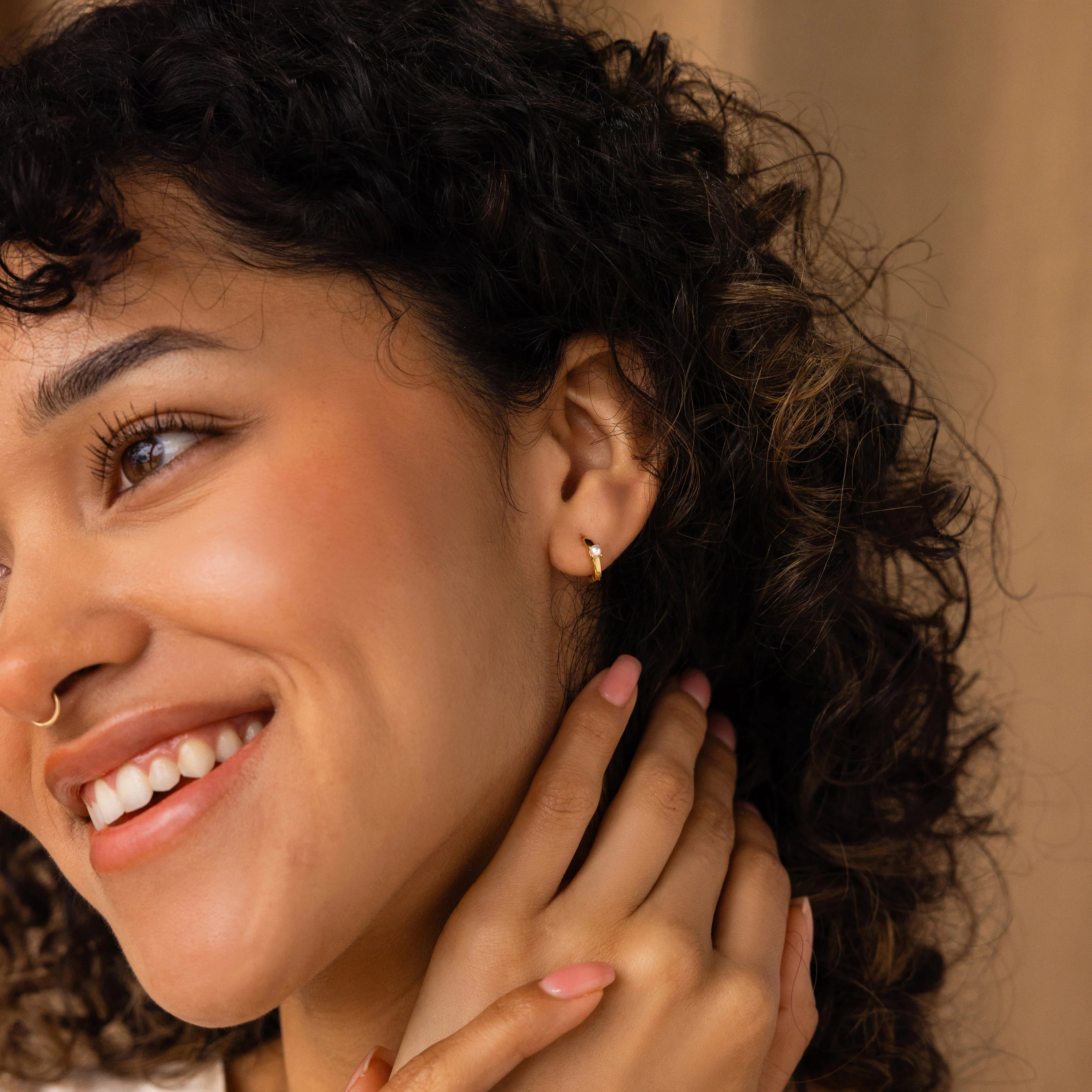 Smiling woman with curly hair, wearing a nose ring, Tiny Pearl Huggies, and light pink nail polish.