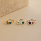 The Duo Heart Birthstone Ring and two other delicate heart-shaped gemstone rings in different colors are displayed on a beige surface.