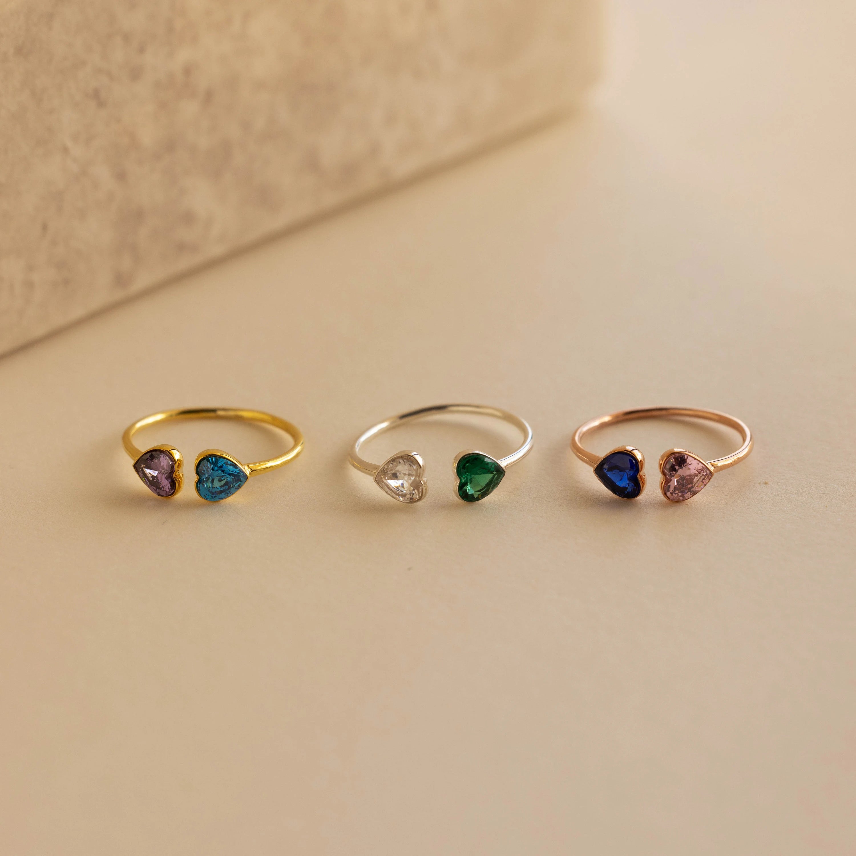 The Duo Heart Birthstone Ring and two other delicate heart-shaped gemstone rings in different colors are displayed on a beige surface.
